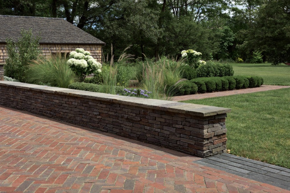 A red brick terrace laid in herringbone meets a low stacked-stone seat wall with a flat stone cap, with hydrangeas, grasses and clipped shrubs planted behind and a lawn to the right