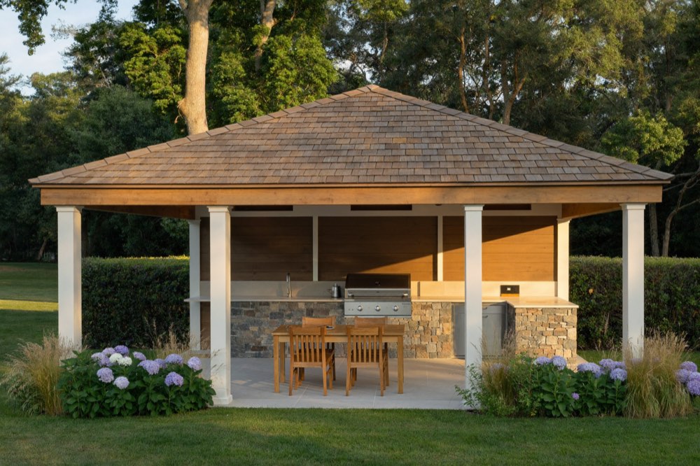 An open cabana with a shingled hip roof on white posts, sheltering a stone-clad outdoor kitchen counter with a built-in grill and a wooden dining table, framed by blue hydrangeas and grasses