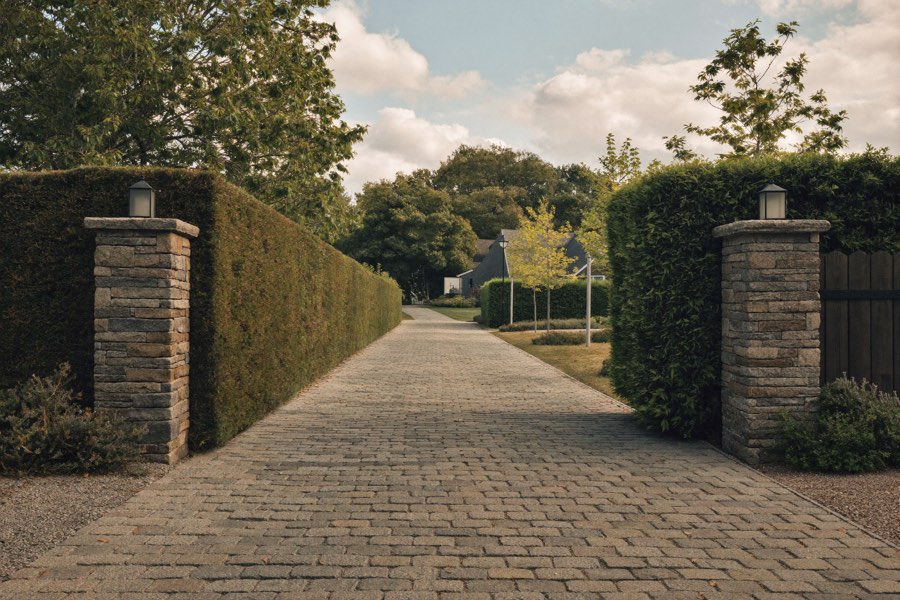A cobblestone driveway entering between two stacked-stone pillars topped with lanterns and tall clipped hedges, leading toward a shingled house under trees