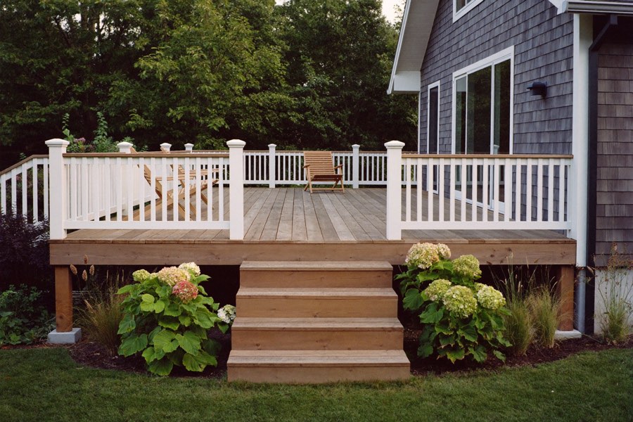A raised wood deck with white railings at the back of a grey shingled house, with wide wooden steps down to the lawn between hydrangeas