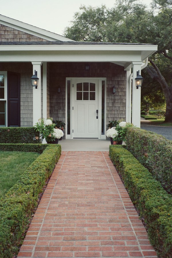 A straight red brick walk between low clipped hedges leading to a white front door on a shingled porch with lanterns and potted white hydrangeas