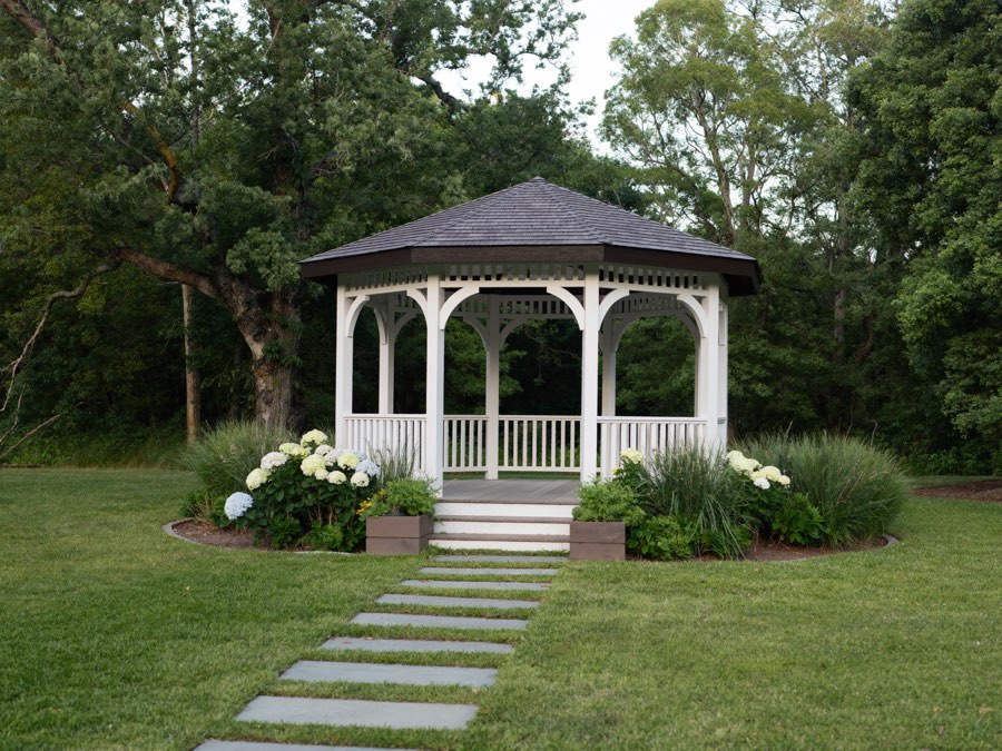 A white octagonal gazebo with a dark shingled roof at the edge of a lawn, reached by flat stepping stones and surrounded by white hydrangeas and grasses