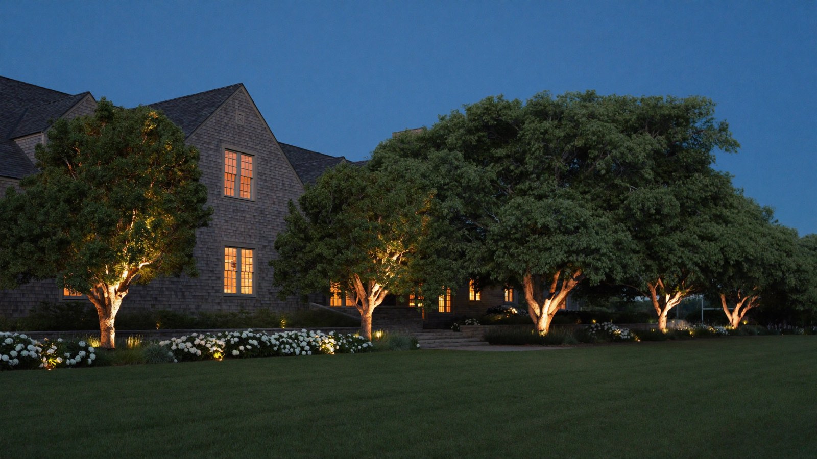 A large shingled house at blue hour with warm lights shining up into a row of mature trees, white hydrangeas along a terrace and a wide dark lawn in front