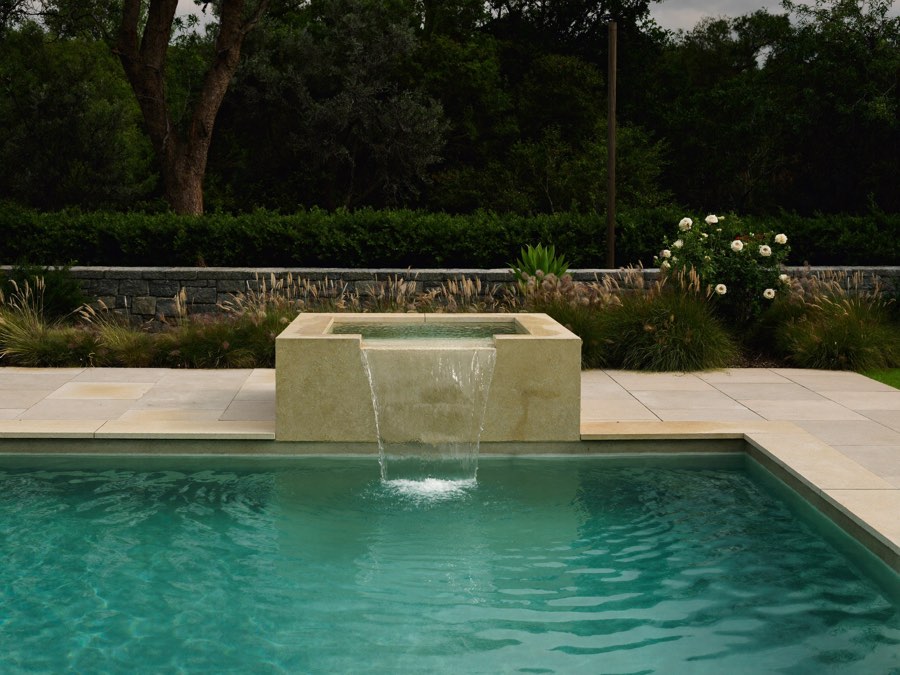 A raised square spa with pale stone walls spilling a sheet of water into a turquoise pool, with pale stone paving, a low stone wall, grasses and white roses behind