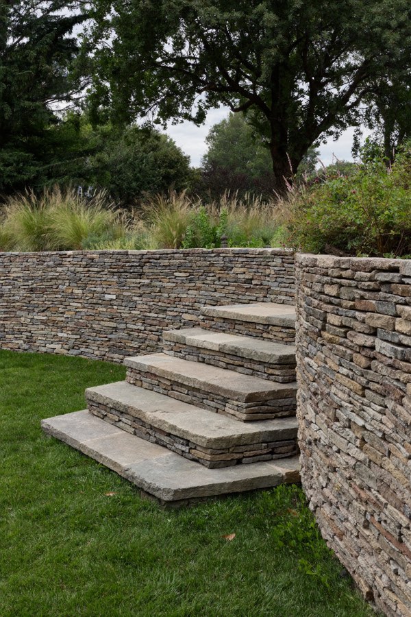 Broad stone steps rising between sections of a curved dry-stacked stone wall, with grasses and shrubs planted along the top and a lawn below